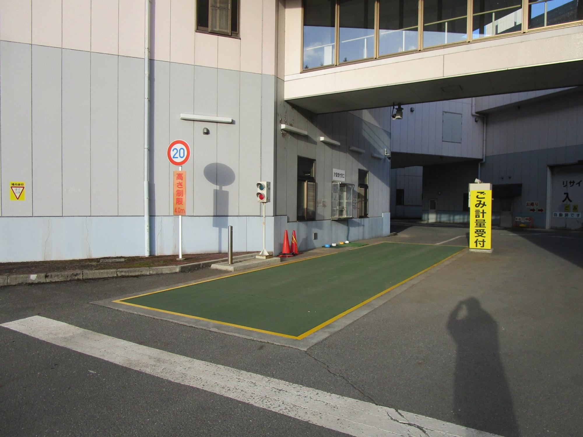 地面に設置された緑色の計量機に黄色い枠線が引かれ、「ごみ計量受付」と書かれた看板が立ててある計量機を斜めから撮影した写真