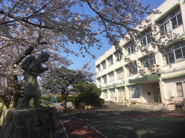 左側に二宮金次郎像と桜の花、右側にマキノ東小学校の校舎が見える写真