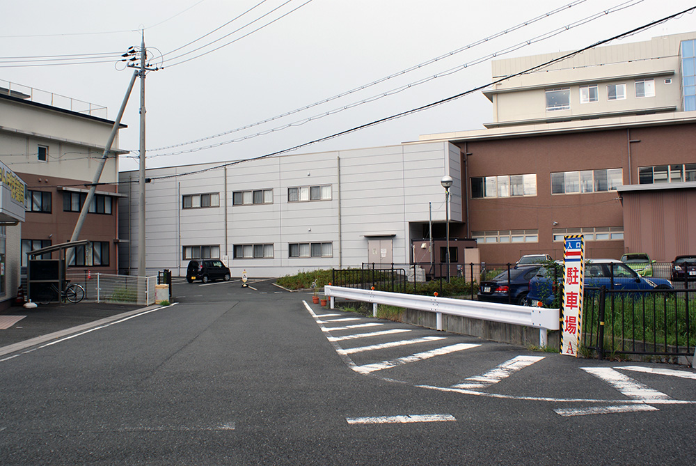 正面奥に茶色や灰色のコンクリート建物が建っており、右側のガードレール横に「入り口 駐車場A」と書かれた看板が置かれている写真