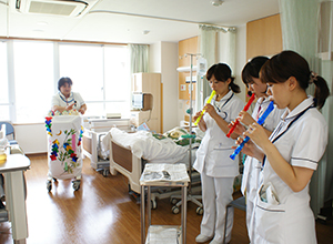 病室で、黄色、赤色、青色の縦笛を吹いている白衣を着た看護部の女性3名と、白い布に折り紙がついたキャスター付きワゴンを押している看護部の女性1名の写真