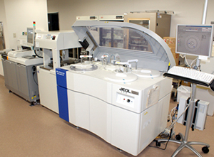 操作用のモニターとキーボードの横にカバーの開いた大きな生化学分析装置が置かれている写真