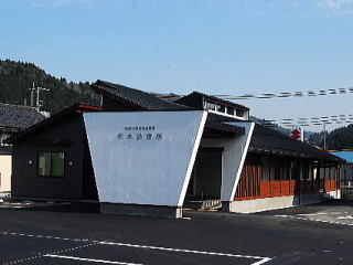 逆三角形のような形の白い壁に朽木診療所と書かれその奥に1階建ての建物が続く高島市民病院朽木診療所の写真
