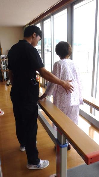 平行棒で歩行練習をしている女性と介助を行っている理学療法士の男性を後方から写した写真