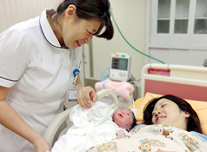 産まれたばかりの赤ちゃんと母親がベットに横たわり、看護師がベットの横で笑顔で赤ちゃんを見ている写真