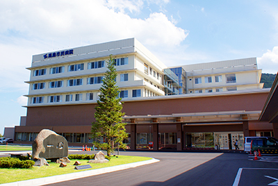 茶色と白色の大きなコンクリート建物があり、左側の芝生に石碑や1本の木が立っている高島市民病院を正面に見た写真