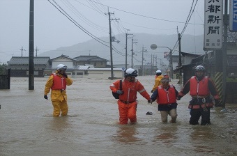 大雨で水が膝の高さまで上がった道路を消防団員が女性の両手を握って避難を行っている水害時の様子の写真