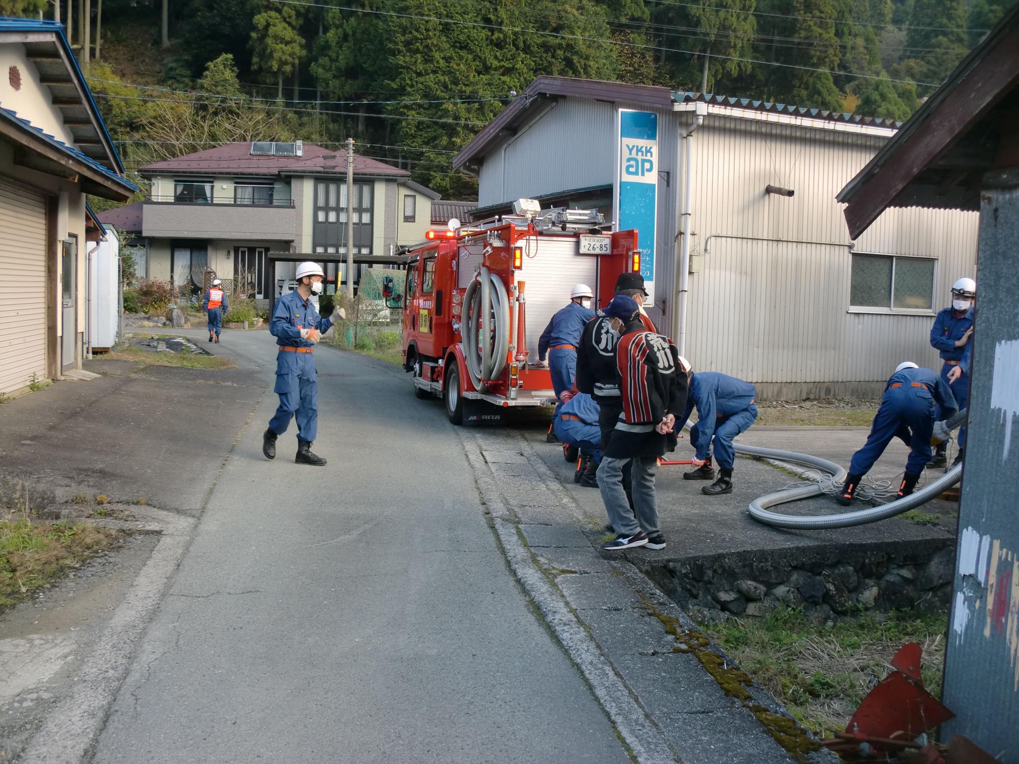 細い路地の端に消防車を停車させ消防団員がホースを持っている訓練の様子の写真
