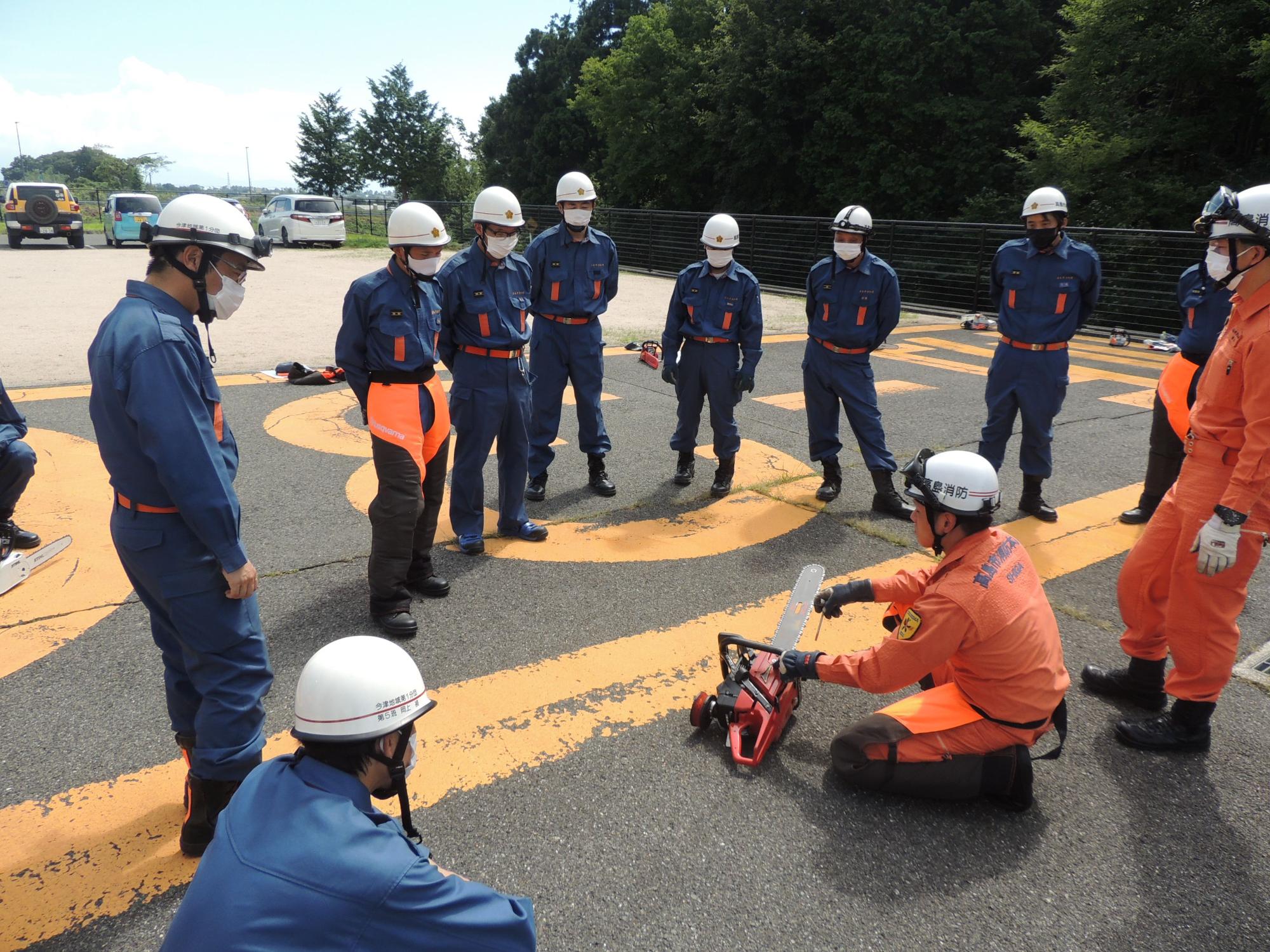 オレンジ色の作業着を着用した先輩の消防団員がチェーンソーのメンテナンスを行っている手本を講習会に参加した消防団員が見ている写真