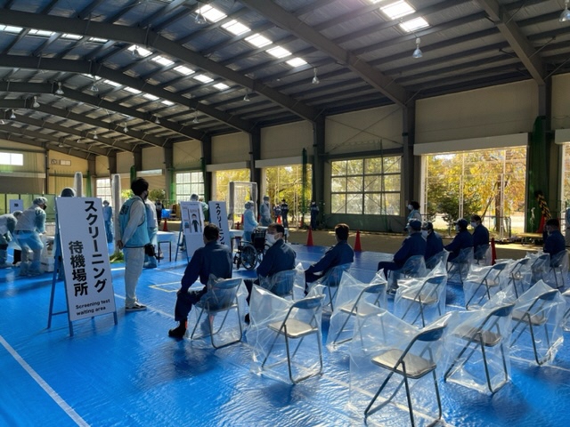 スクリーニング待機場所と書かれた立て看板の手前に設置された透明のビニール袋で覆われたパイプ椅子に消防団員が座っている写真