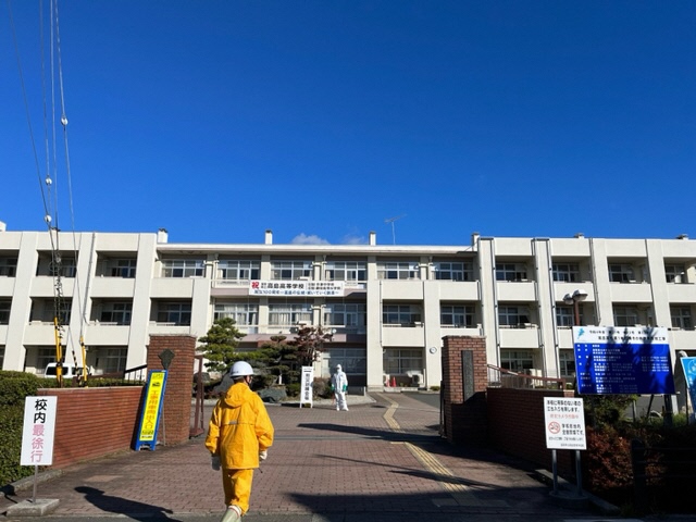 高島高校の校門の先に防護服を着た消防団員が立ち、黄色の雨具を着た消防団員が校門に向かって歩いている写真
