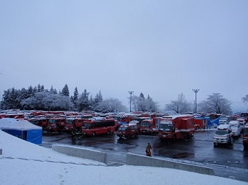 雪が積もるなか大きな駐車場にたくさんの消防車や救急車が停められている写真