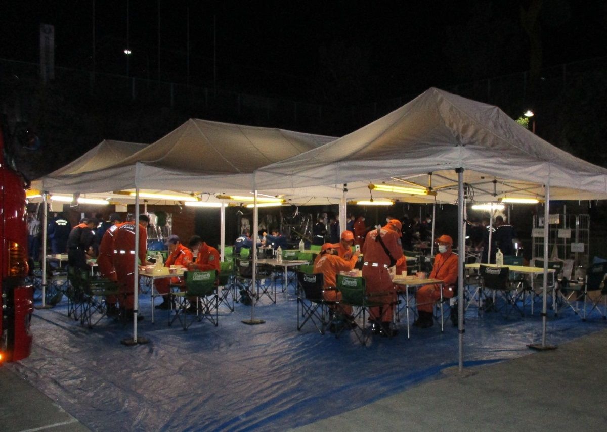 テーブルと椅子が設置された大きなテントの下でオレンジ色の作業着を着用した消防団員が座っている野営訓練の様子の写真