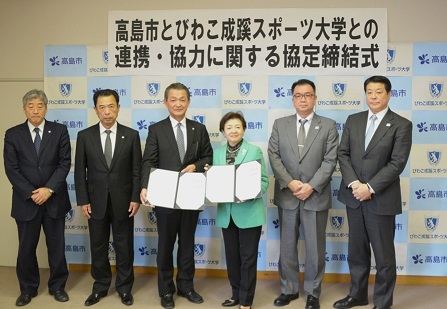 協定締結式の会場で、左右に2人ずつ関係者が立ち、中央に協定書を持った福井市長と嘉田学長が立っている写真