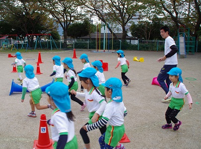 青い帽子をかぶった園児たちがカラーコーンが置かれている運動場で、大人の指導を受けながら運動をしている様子の写真