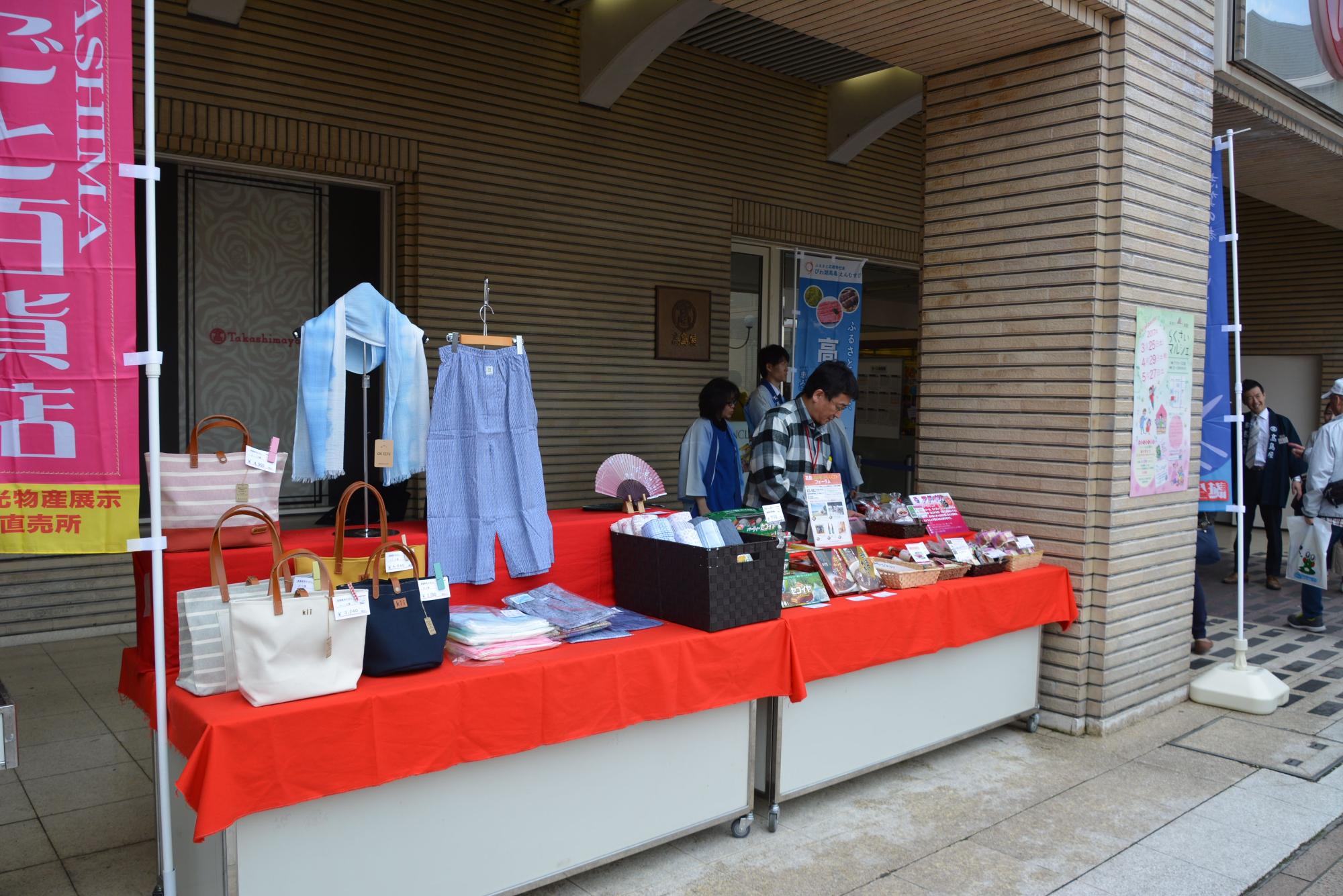 会場の入り口横に、カバンや衣類などの特産品が並べられた「高島市物産フェア」のお店の写真
