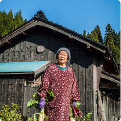 収穫した大根を両手に持ち小屋の前に立っている澤井キミコさんの写真
