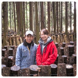 シイタケの原木がたくさん置かれている農園の中で撮影された水口さん夫妻の写真