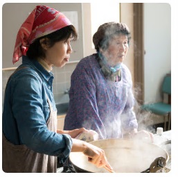 湯気のあがる大きな鍋をかき混ぜ、調理をする女性2人の写真