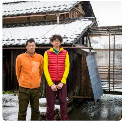 屋根や地面に雪が積もる小屋の前に立つ2人の男性の写真