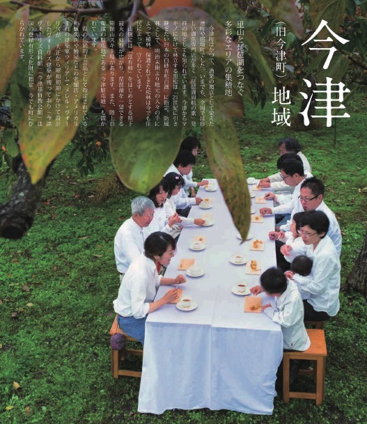 10人ほどの老若男女が白い服を着て柿畑に置かれた長机に座り柿を食べている写真