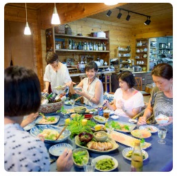 青いテーブルクロスが敷かれたテーブルにたくさんの料理が並び料理教室の参加者が席についている写真