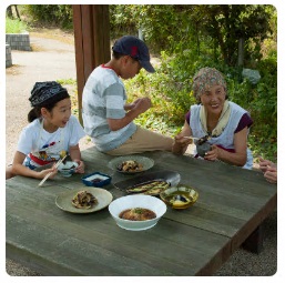 外に置かれた木の台に数種類の茄子料理が並び、白崎さんとお孫さんが料理を食べている写真