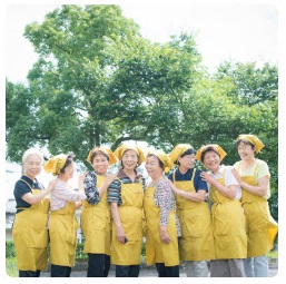 お揃いの黄色いエプロンと三角巾を付けた郷土料理伝承の会の方々が笑顔で写る集合写真