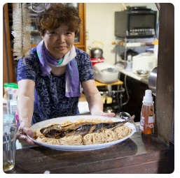 大皿に乗せられた麺の上に焼き鯖が丸ごと乗った「鯖そうめん」を持っている海老澤文代さんの写真