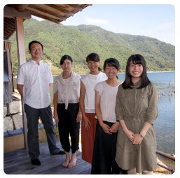 奥に湖や山が見える「湖里庵」の縁側に立つ4人の女子高校生と店主の左嵜さんの写真