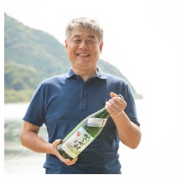 湖を背景にお酒の瓶をもって笑顔を見せる吉田さんの写真