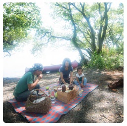湖のほとりにある木陰にレジャーシートを敷き、ランチを楽しむ家族3人の写真