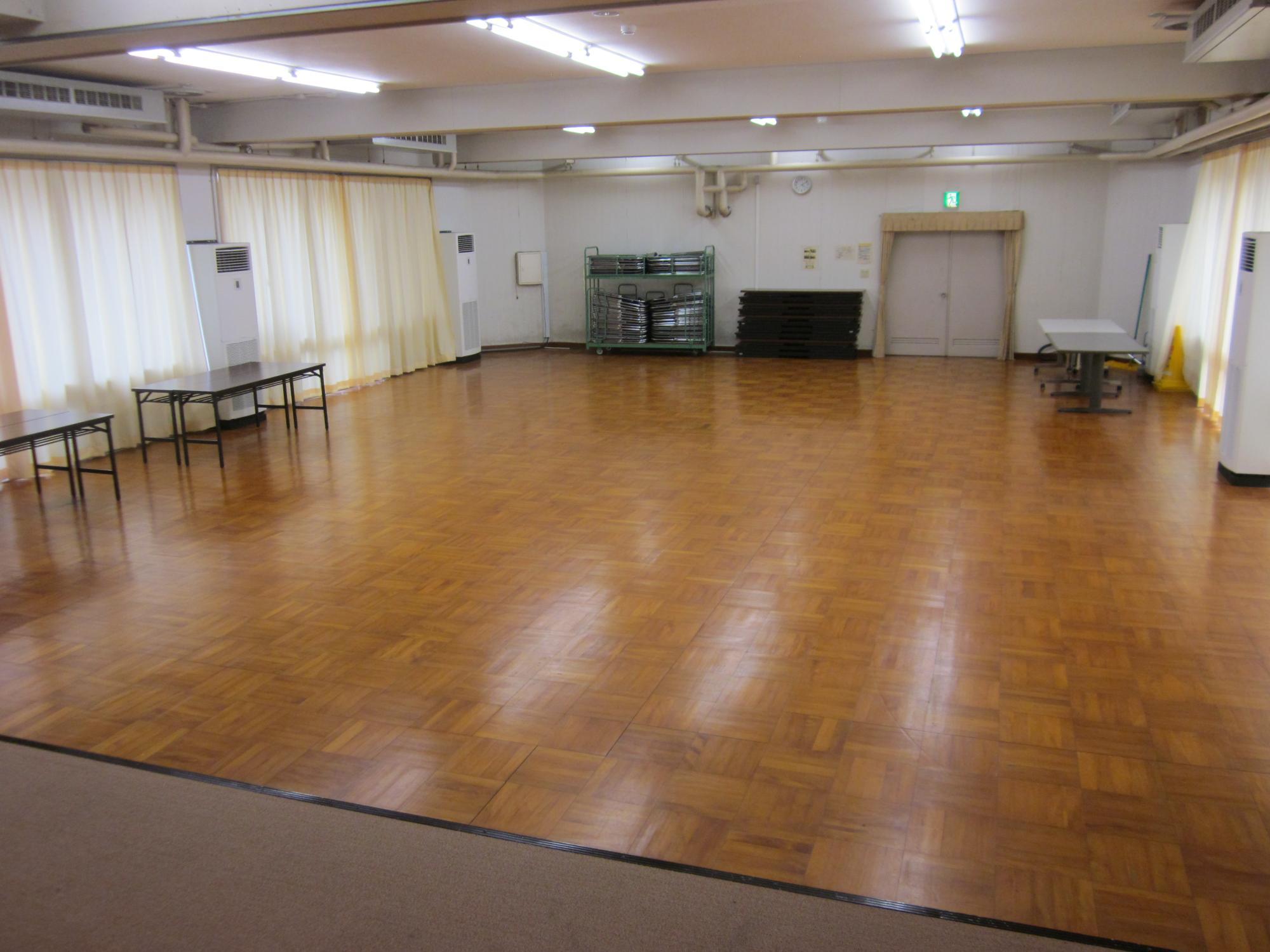 床は木のフロアーの小ホール、手前がステージ、左サイドに会議机、奥にパイプ椅子、その右が出入り口