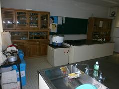 前方には黒板と食器棚が設置され、やかん、ガス窯、調理スペースのある調理室の写真