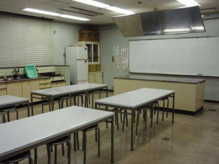 テーブルと椅子、冷蔵庫や調理台があり前方には大きなホワイトボードが設置された調理実習室の写真