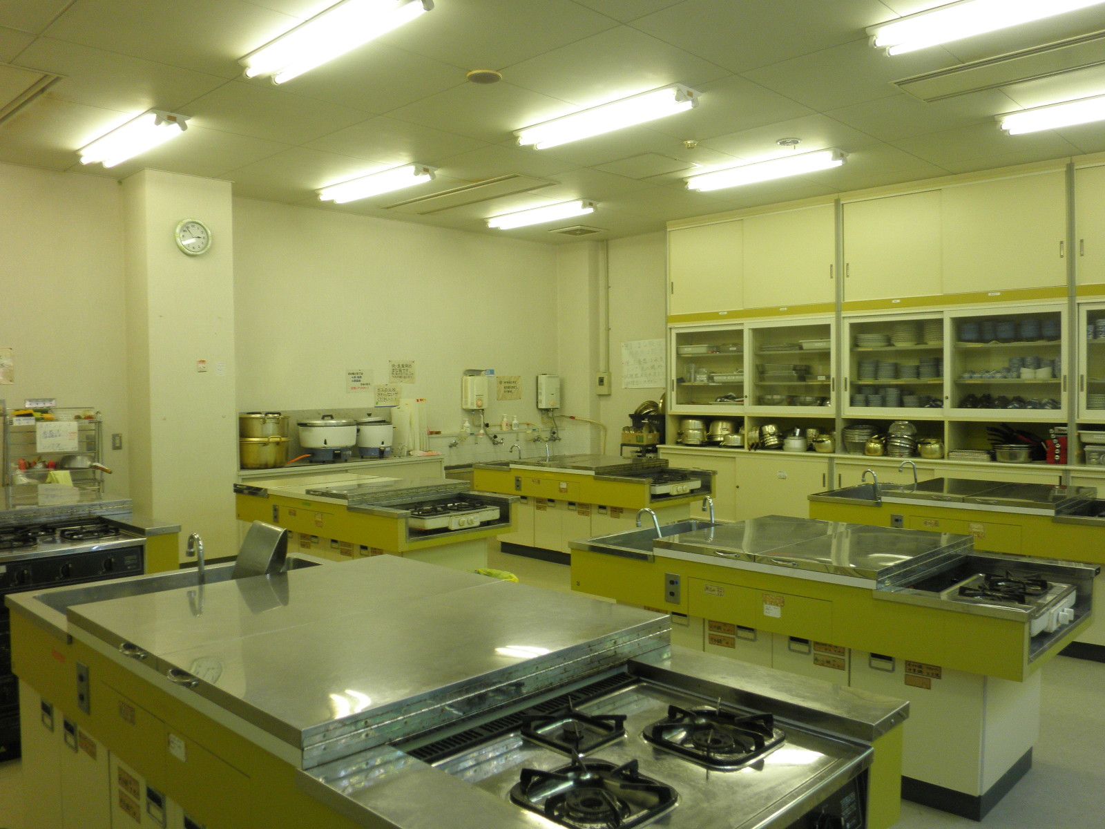 右側の壁には鍋や調理器具が収納されている棚、ガスコンロが設置されている調理台が6台並んでいる調理室の写真