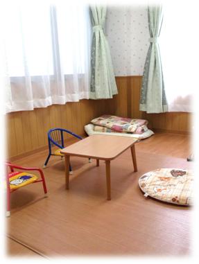 レースのカーテンが閉められ、端に畳んだ布団があり、テーブルと子ども用の椅子がある室内の写真