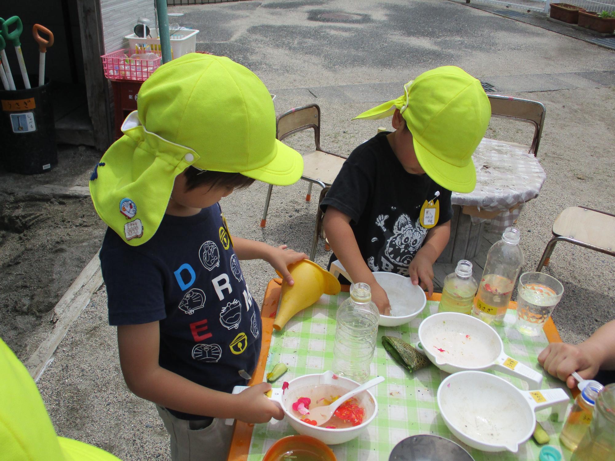 黄色の帽子をかぶった園児2人が容器に入った色水で遊んでいる写真