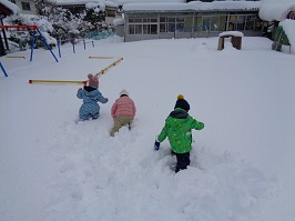 後ろの園舎や木にも雪が積もり、園庭の雪の上で遊ぶ子供達の写真