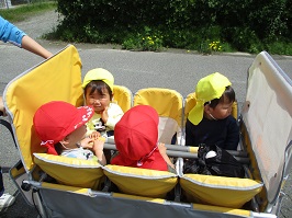 6人乗りのお散歩カートに赤い帽子をかぶった園児2名と黄色の帽子をかぶった園児2名が乗って散歩している写真