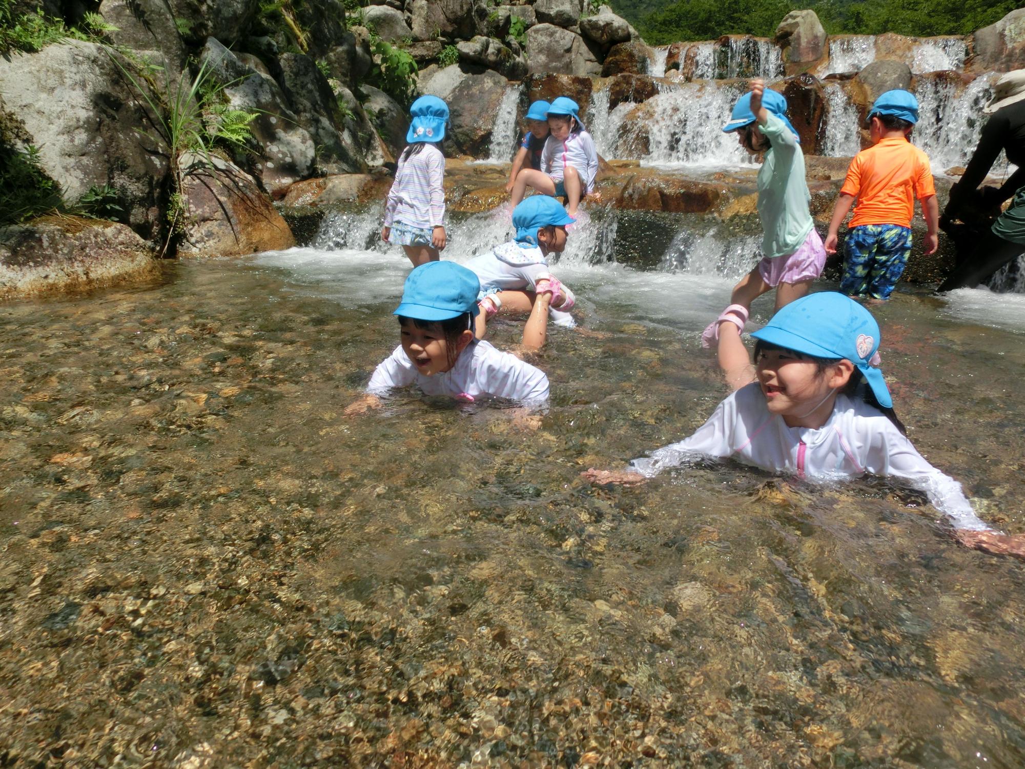 後ろには小さな滝があり、水色の帽子をかぶった園児達が川遊びをしている様子の写真