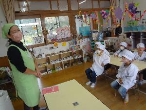 給食の割烹着、給食帽をかぶって椅子に座り、食育サポーターの話を聞いている園児たち