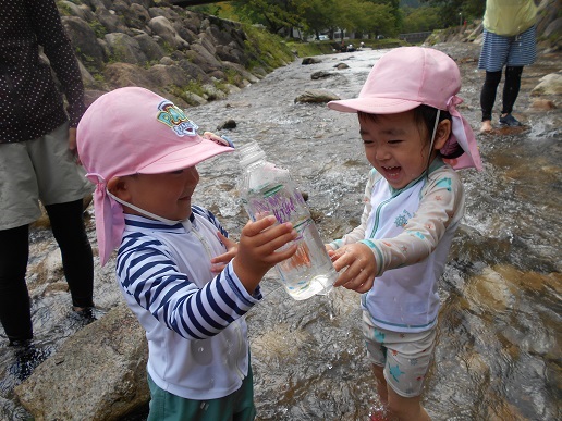 水着姿の子ども2人がペットボトルに入れた水で川遊びをしている
