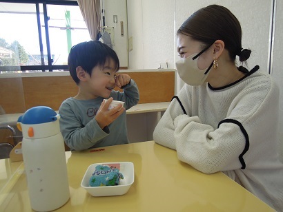 親子が向かい合ってすわり、おやつを食べている子どもに優しく微笑みかけている母親