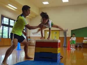 体操服姿の園児が跳び箱を飛ぶのを、大きく足を広げて補助している運動遊びコーチ