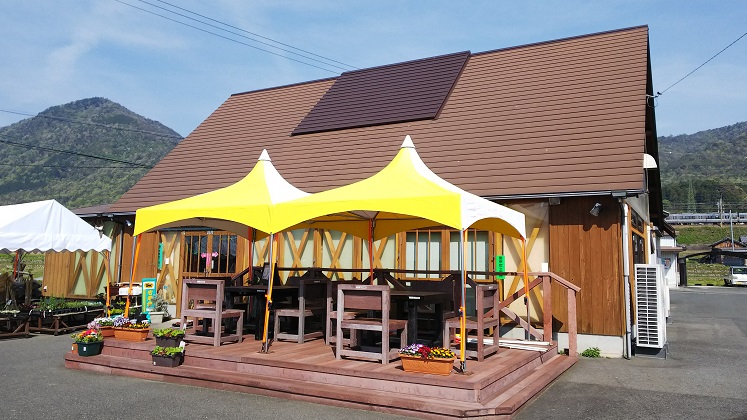 木材を基調とした三角屋根の建物の前に、外で食事ができるよう黄色いテントの中にテーブルと椅子が設置されている写真