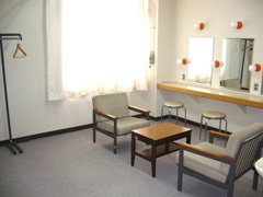 中央に椅子が2脚セットされた小さなテーブル、右側の壁に鏡が設置され丸椅子が並んでいる楽屋の写真