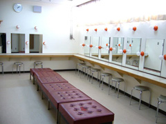 中央に6脚正方形の椅子が並び、奥と右側の壁に鏡ごとに丸椅子がそれぞれ並んでいる出演者控室の写真