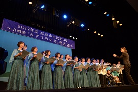 水色のドレスを着た来た女性たちと私服姿の若い人たちがステージに立って歌を歌っているシーンの写真（高島市市民劇2016「みずうみの詩」ページへリンク）