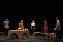 テーブルと椅子が並んでいるステージで5名の人たちが間隔をあけて立って話をしているシーンの写真