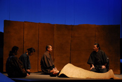 体にゴザをかけられた横たわっている男性のそばで向かい合って座っている2名の男性の会話を2名の女性が左側に並んで座って聞いているシーンの写真（高島市市民劇2010「琵琶湖治水の物語」ページへリンク）